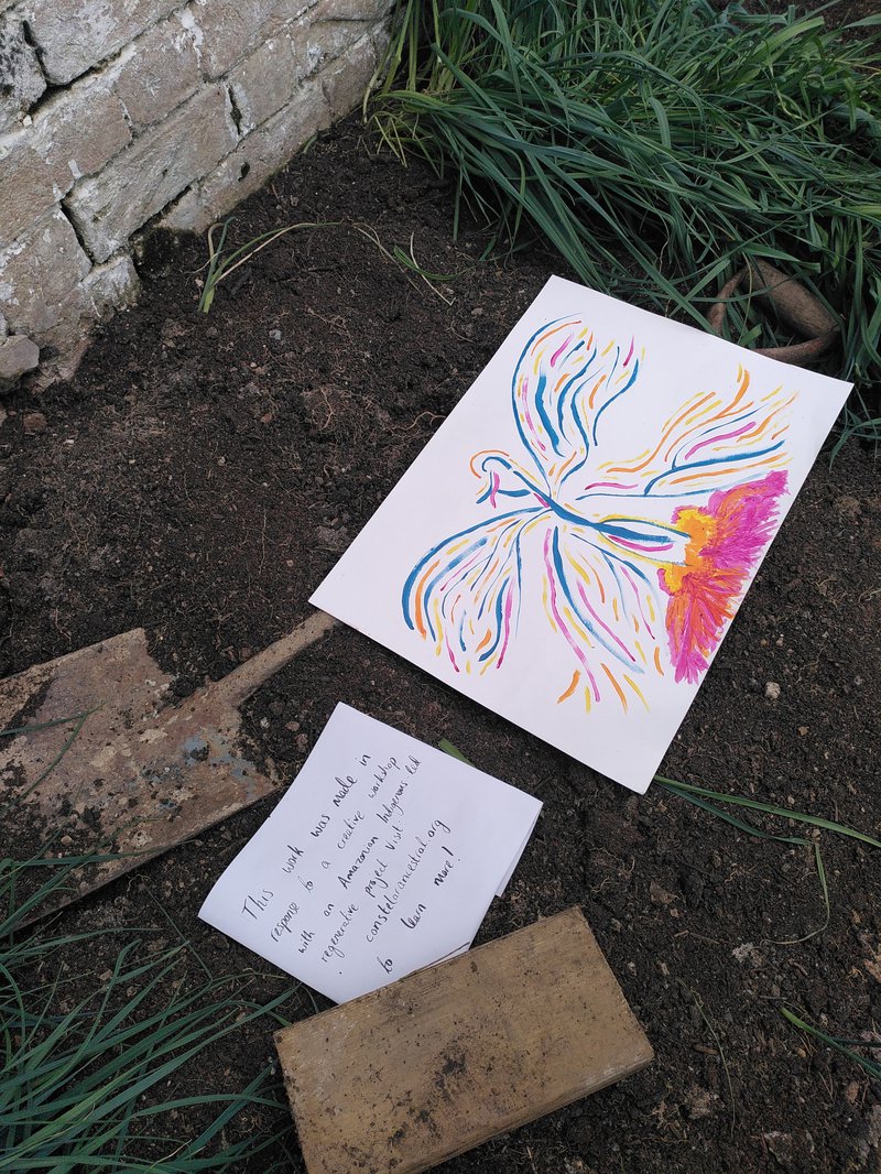 Photo of a painting of an abstract bird, lying on the soil above a spade in the old greenhouse