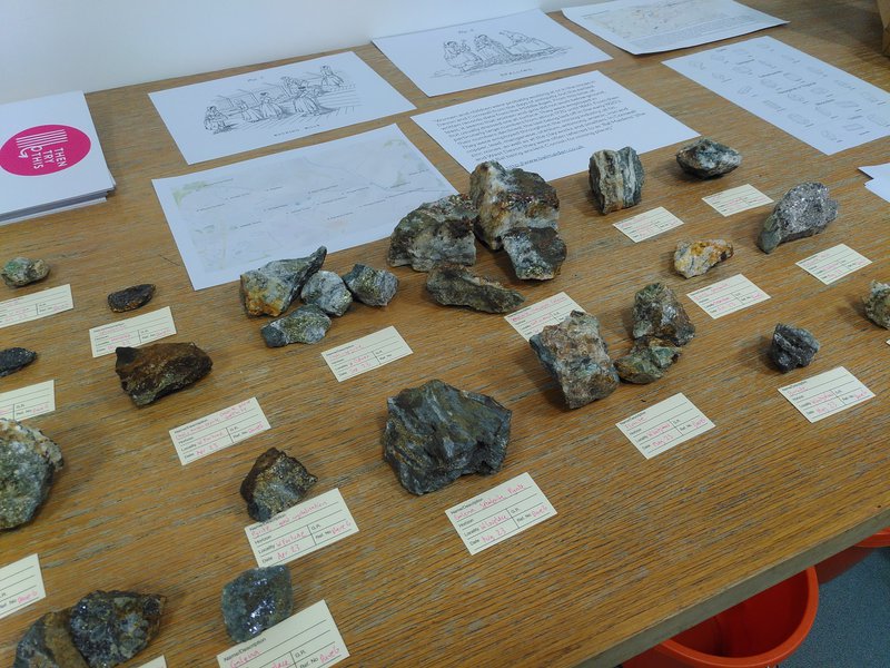 A small collection of rocks with small labels next to them explaining what they are and where they come from. In the background is information on the Bal Maidens and maps of mine sites in Gwennap.