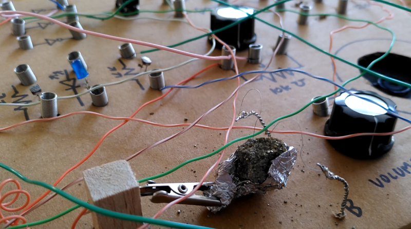 Closeup of a cardboard box with compoents, dials and wires. A lump of crystalline pyrite is shown held by a crocodile clip and a pair of small wires connect it to a circuit.