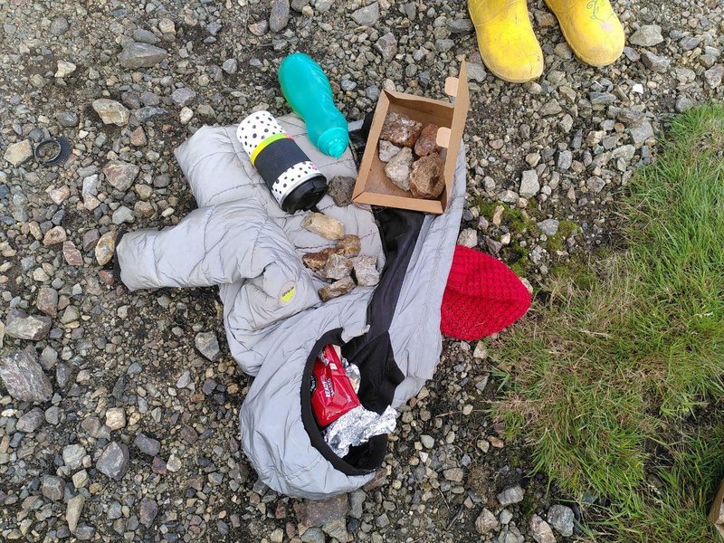 A small collection of rocks in a cardboard box on top of a childs coat with a pack of biscuits. Two small yellow wellies visible at the top of the image.