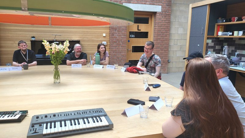 Seven people sat chatting and looking amused, around a very large round wood table, with flowers in the middle, and a couple of keyboard synths.