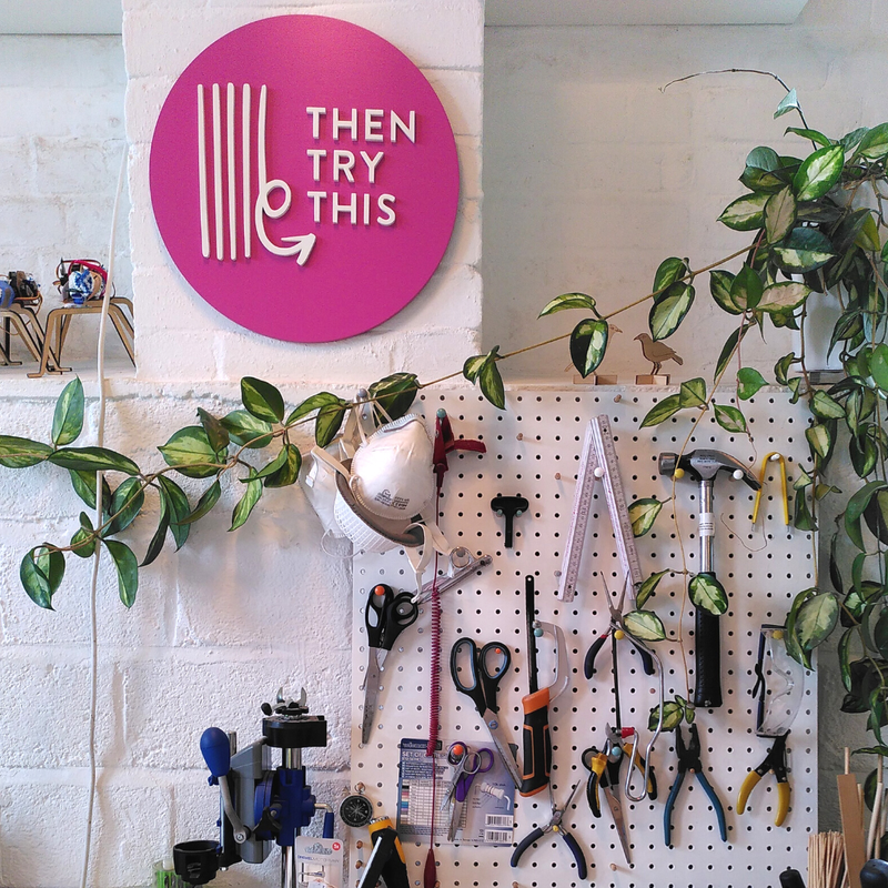Photo of our logo on a round pink sign, with a peg board full of tools below it, and a hoya plant tangled around