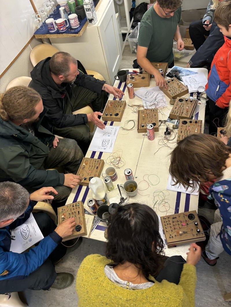 A table viewed from above with lots of cardboard synths being wired together by people.