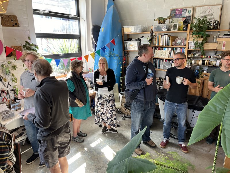 Seven people standing in a studio decorated with bunting, with bookshelves and kayaks in the background, plants in the foreground.