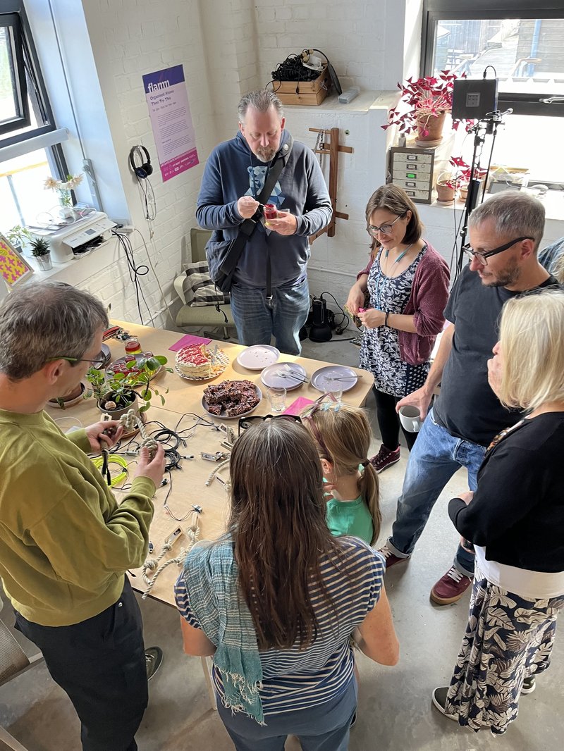 Seven people from above surrounding a desk with cake and experimental electonic interfaces on.