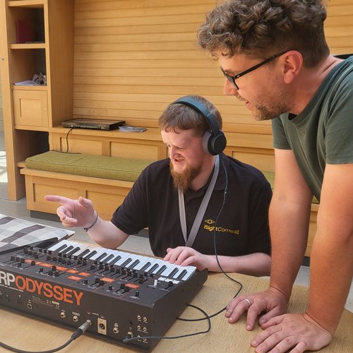 A person playing an ARP ODYSSEY synth, wearing headphones, one hand on the keyboard and another pointing forward, and anther person leaning on the table next to them watching and smiling