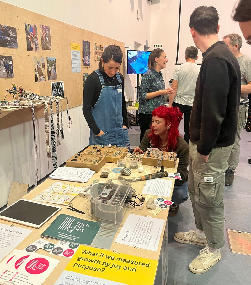 A group of people around a table with diy electronics, a rock hammer, air quality sensor on. Pictures on the board on the wall to one side.