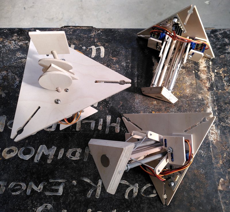 Photo of three virus modules, each consists of an outer wood triangle and a smaller inner wood triangle containing a magnet - between them are metal poles and wood poles that move up and down, driven by four servos and a small circuit board.