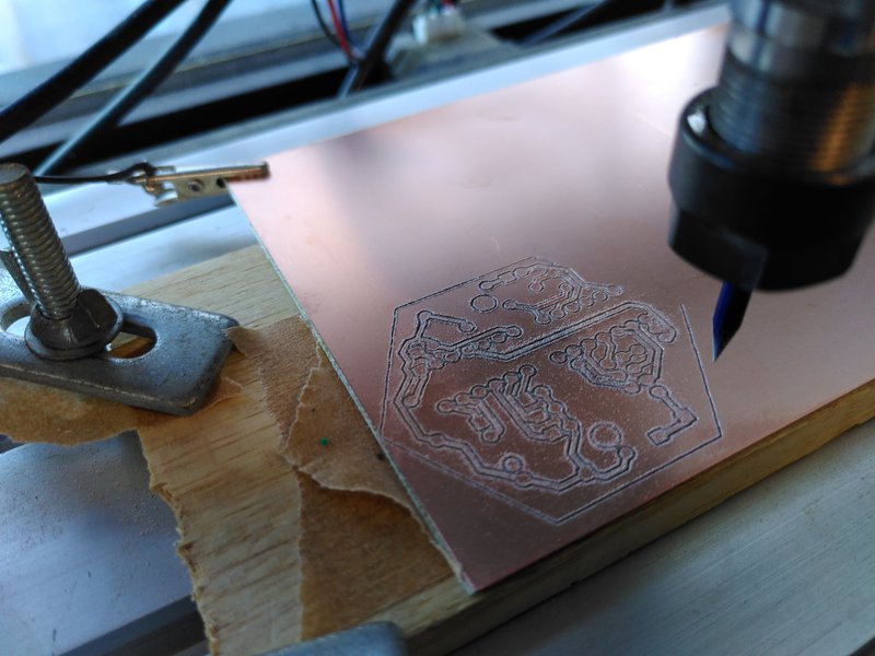 Circuit board being milled in the CNC machine, raw copper clad board being cut by a small V shaped bit, clamped to the base plate of the CNC