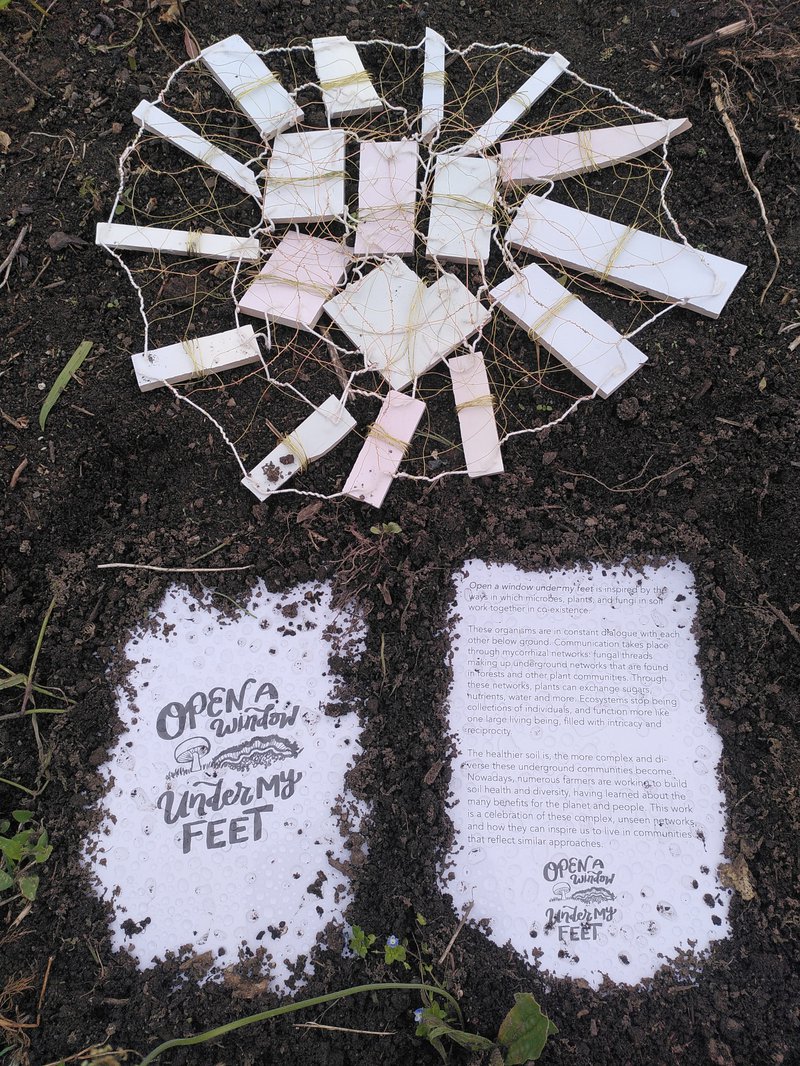 Photo of sign saying open a window under my feet, embedded in the soil, and another sign with a lot of text on it, together with some tile offcuts wired together into a network