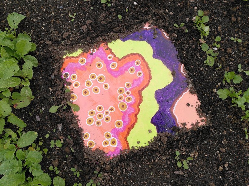 photo of a tile painted like bacterial growth, embedded in the soil between seedlings