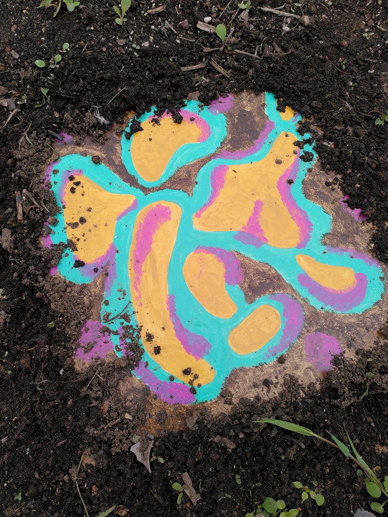 photo of a tile painted like bacterial growth, embedded in the soil between seedlings
