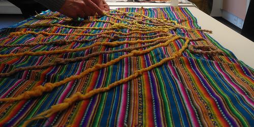 A knotted string khipu attached to a bright multicolored weaving being handled by someone.