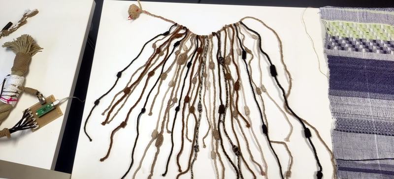 A khipu on a table with a circuit board on the left and a weaving on the right.