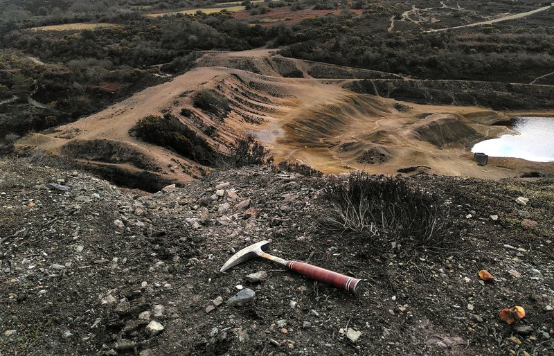 A geological rock hammer on the ground at the top of a hill of mine waste with a view of water and gravel forming the Wheal Maid tailings dam. View from East Wheal Maid mine shaft.