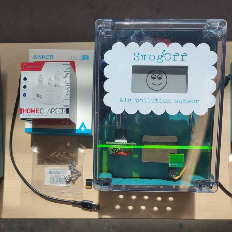 A smogoff sensor, teal coloured box with clear front, fluoro yellow shelf holding sensors, a smiley face on a screen, and a cloud shaped sticker around the screen