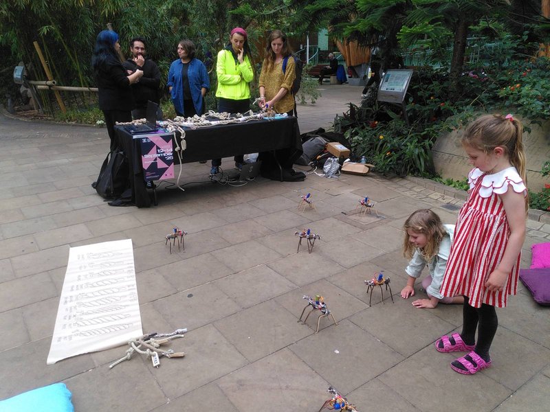 Two children talking to small woven robots while a group of people stand around a table in the background, surrounded by plants.