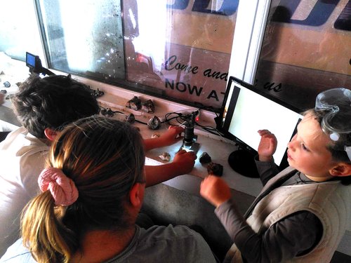 Three people surround a usb microscope attached to a monitor, with rocks on a table.